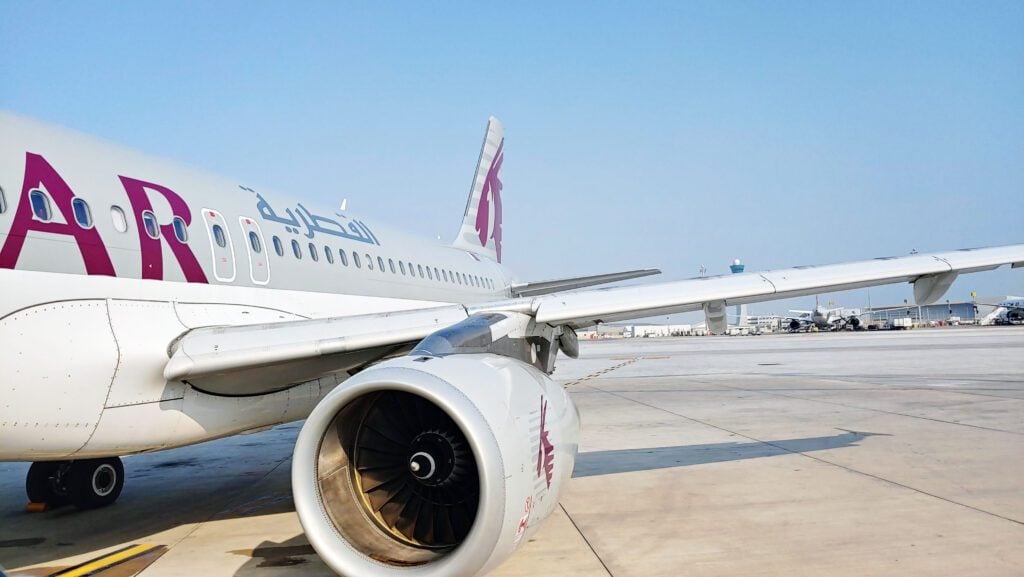 A Qatar Airways airplane is parked on the tarmac at an airport, showing a close-up of its engine, wing, and part of the fuselage under a clear sky.