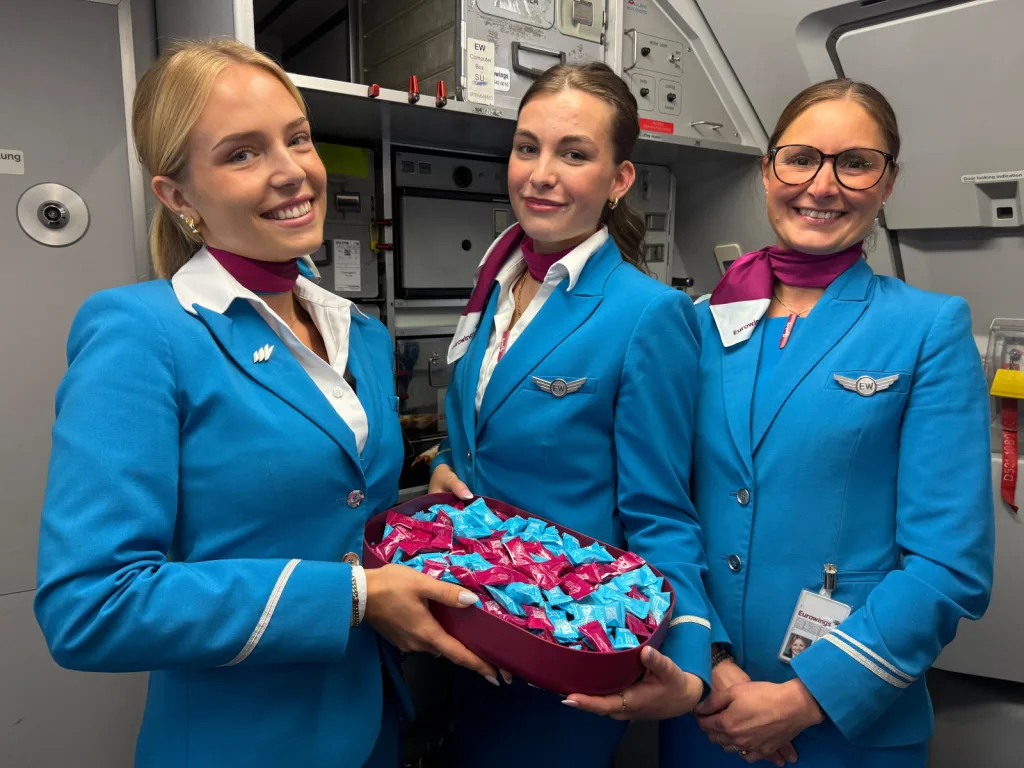 Three flight attendants in blue uniforms stand in an airplane galley, one holding a bowl filled with blue and pink packaged snacks, as Air France adds Daniel Boulud to its US chef roster.