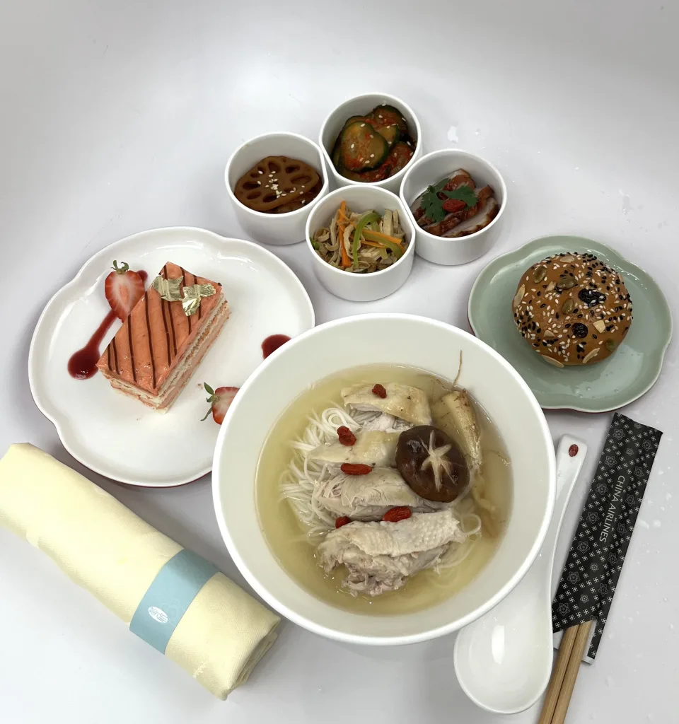 A bowl of chicken and mushroom noodle soup, side dishes, a strawberry dessert, a sesame bun, chopsticks, and a napkin are arranged on a white surface—perfect for sampling after Air France adds Daniel Boulud to its US chef roster.
