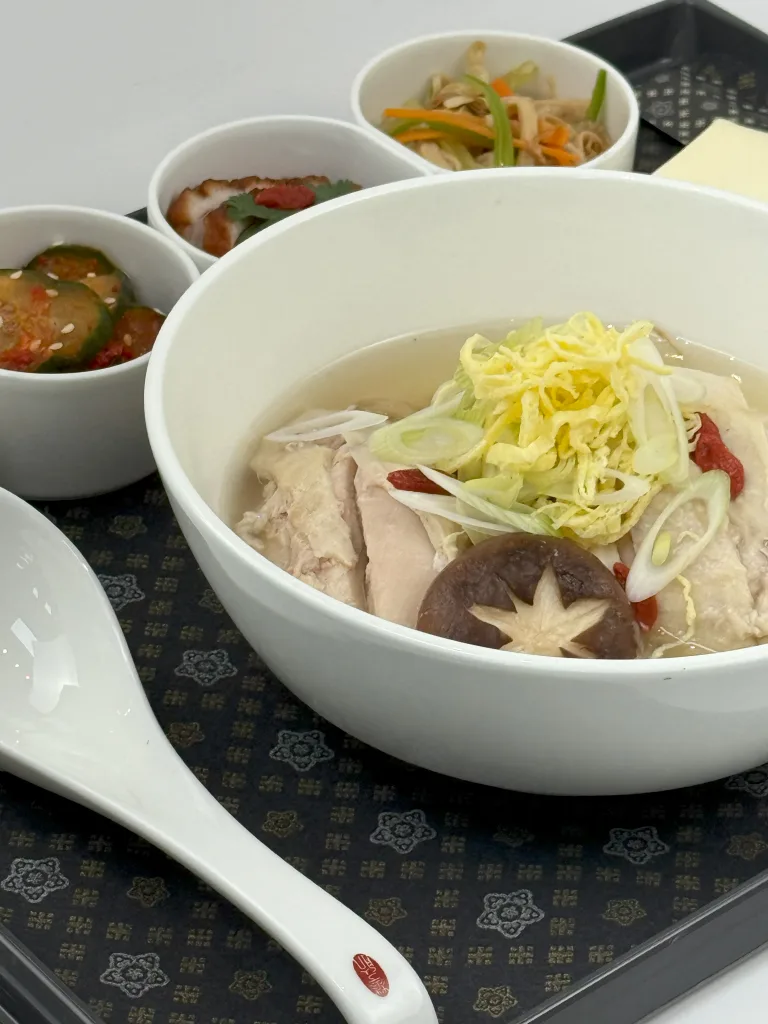 A bowl of chicken soup with mushrooms and shredded vegetables, served alongside pickled cucumber, kimchi, and veggies on a tray with a large white spoon—an experience reminiscent of Air France Adds Daniel Boulud to US Chef Roster.