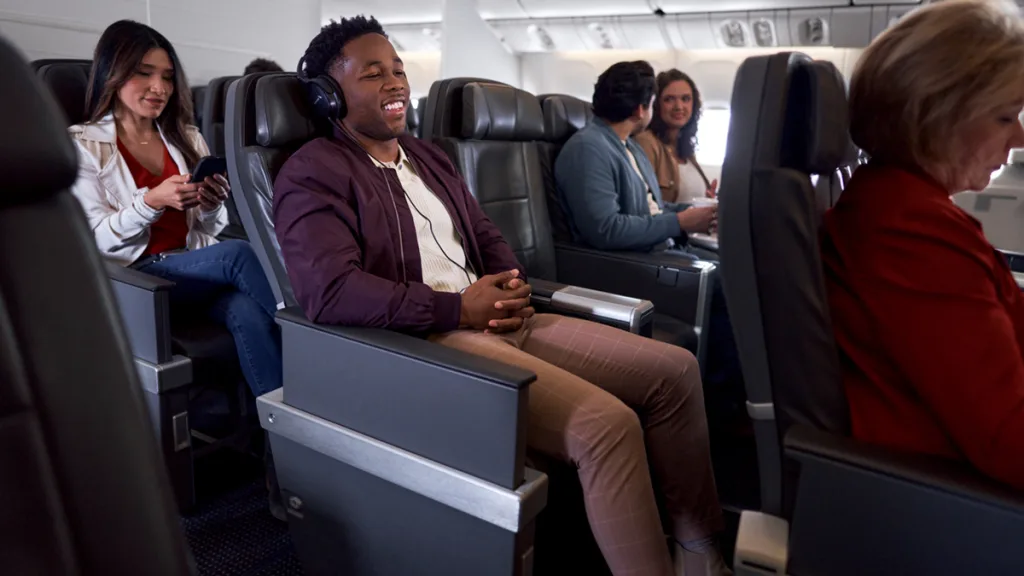 Passengers sit in airplane seats; a man in the foreground wears headphones and smiles, while others enjoy Fiji Airways’ dine on demand service, engage with their phones, or chat with fellow travelers.