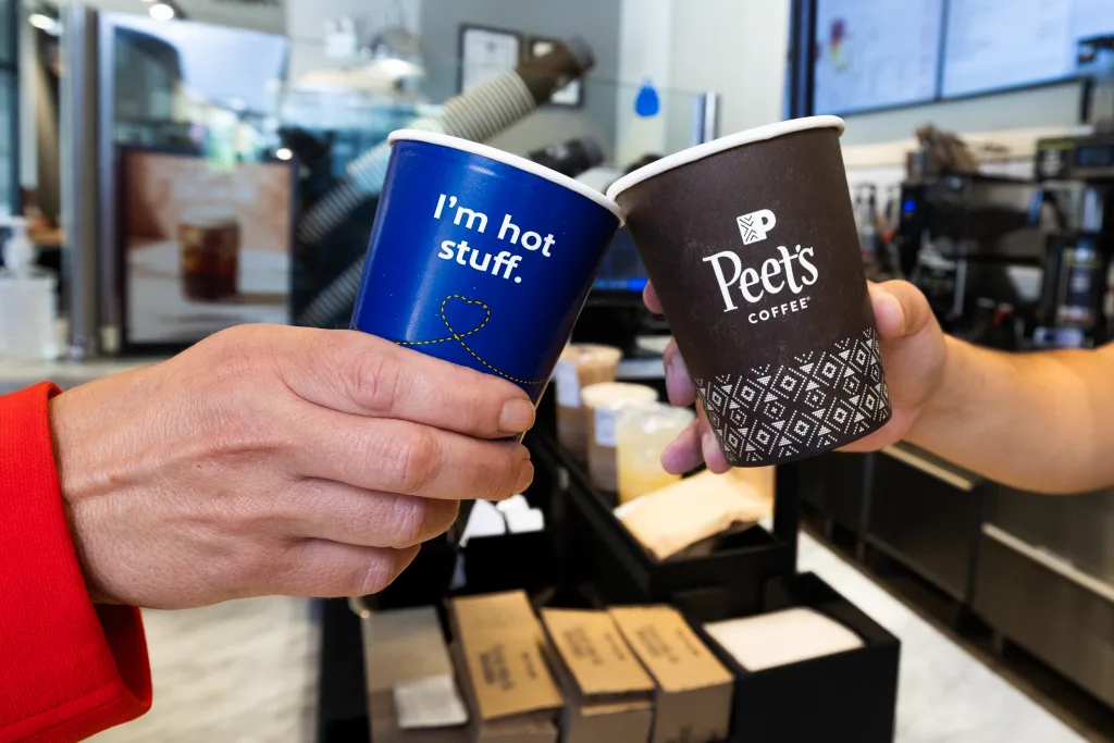 Two people clink coffee cups inside a café—a blue mug reading "I'm hot stuff" and a dark cup with the Peet's Coffee logo—enjoying a moment that rivals aa premium economy transcon comfort.
