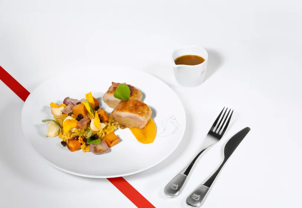 A plated gourmet dish with sliced meat, vegetables, and sauce on a white plate, accompanied by a cup of sauce, knife, and fork on a white surface with a red diagonal line.