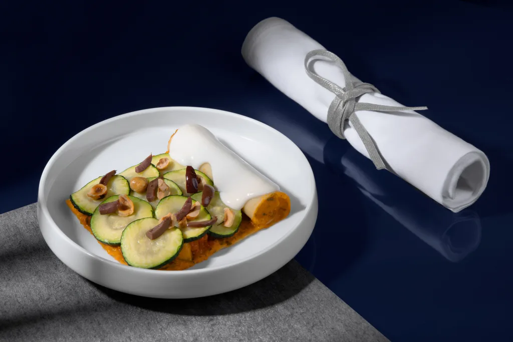 A white plate with sliced zucchini, nuts, and sauce beside a rolled white napkin tied with a gray ribbon on a dark blue surface.