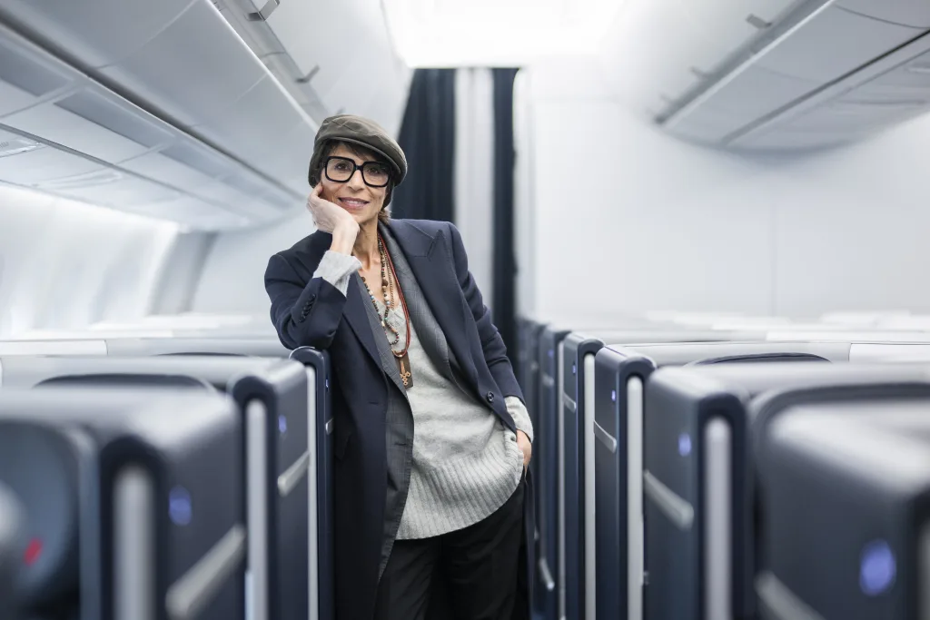 Person wearing a hat, glasses, and layered clothing stands in the aisle of an empty, modern airplane cabin, leaning on a seat and smiling.