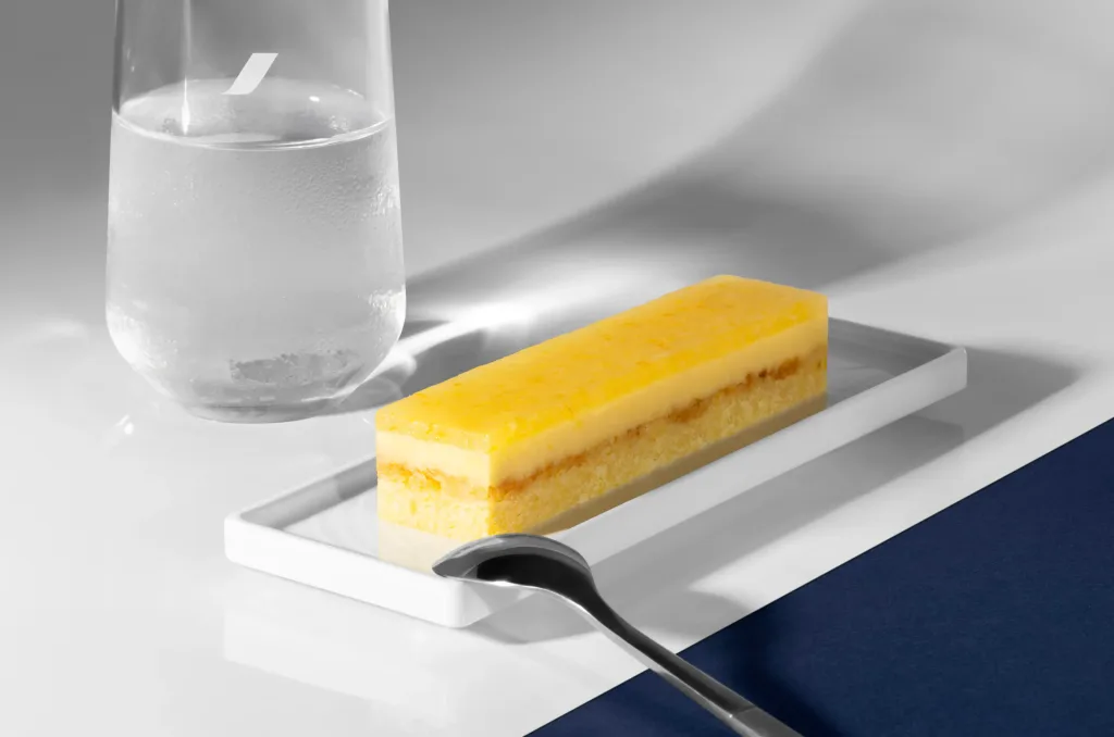 A rectangular slice of layered yellow cake sits on a white tray next to a metal spoon, with a glass of water in the background.