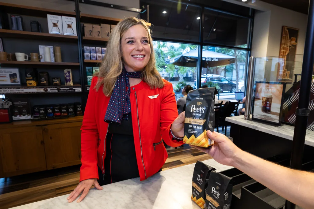 A woman in a red jacket hands a bag of Peet’s Coffee to a customer at a coffee shop counter, where shelves of coffee products evoke the comfort found in AA Premium Economy Transcon service.