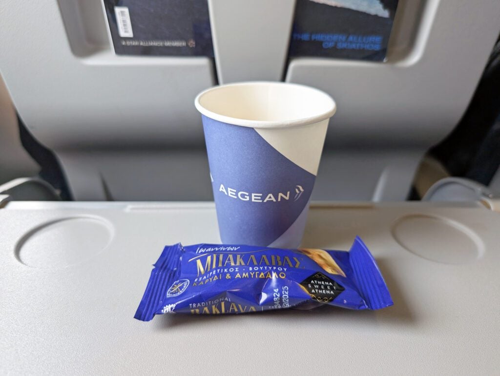 A paper cup with "AEGEAN" branding and a packaged snack rest on an airplane tray table, with seat backs and safety cards visible in the background.