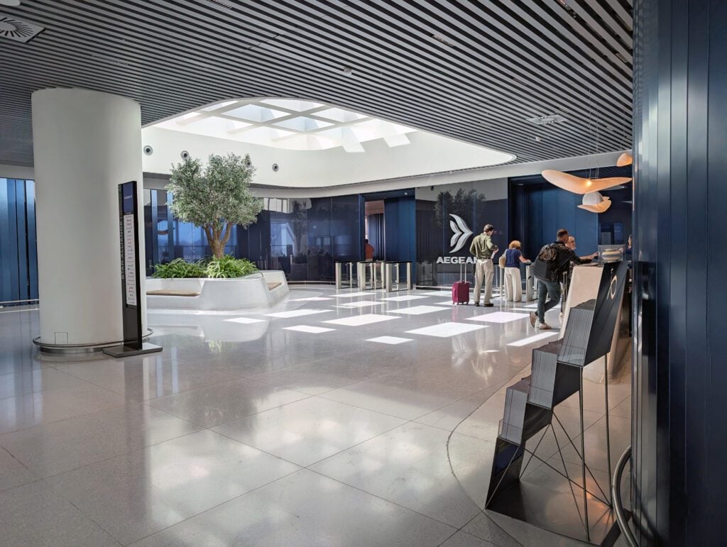 A modern airport lounge with travelers at the check-in counter, an indoor tree, seating area, and Aegean Airlines branding on the wall.