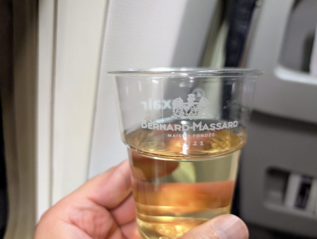 Eine Hand hält in einem Flugzeug in der Nähe eines Fensters einen mit Weißwein gefüllten Plastikbecher mit dem Bernard-Massard-Logo.