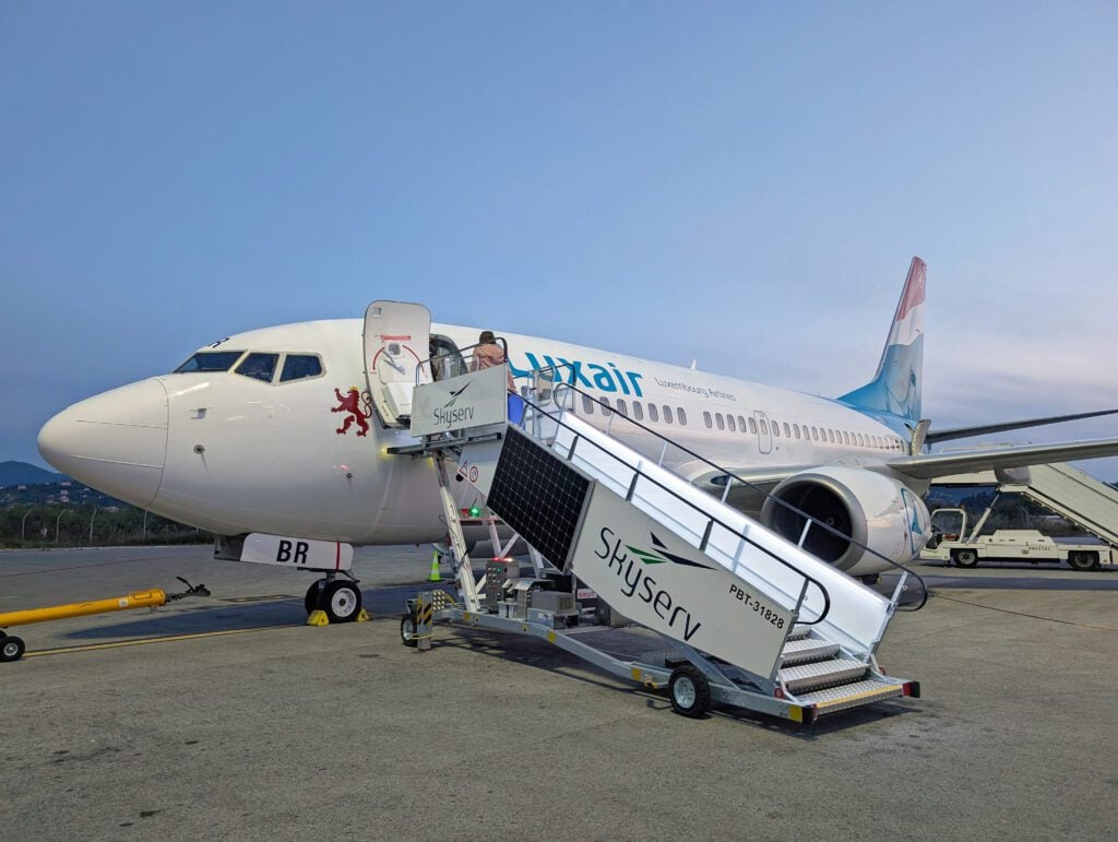 A Luxair passenger airplane is parked on the tarmac at dawn, with stairs labeled "Skyserv" leading to the open aircraft door.