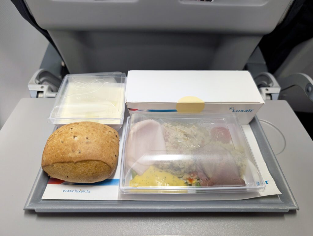 Ein Tablett mit Flugzeugessen der Economy Class mit einem Brötchen, abgepackter Butter und einem abgedeckten Hauptgericht, das auf einem Tabletttisch der Fluggesellschaft steht.