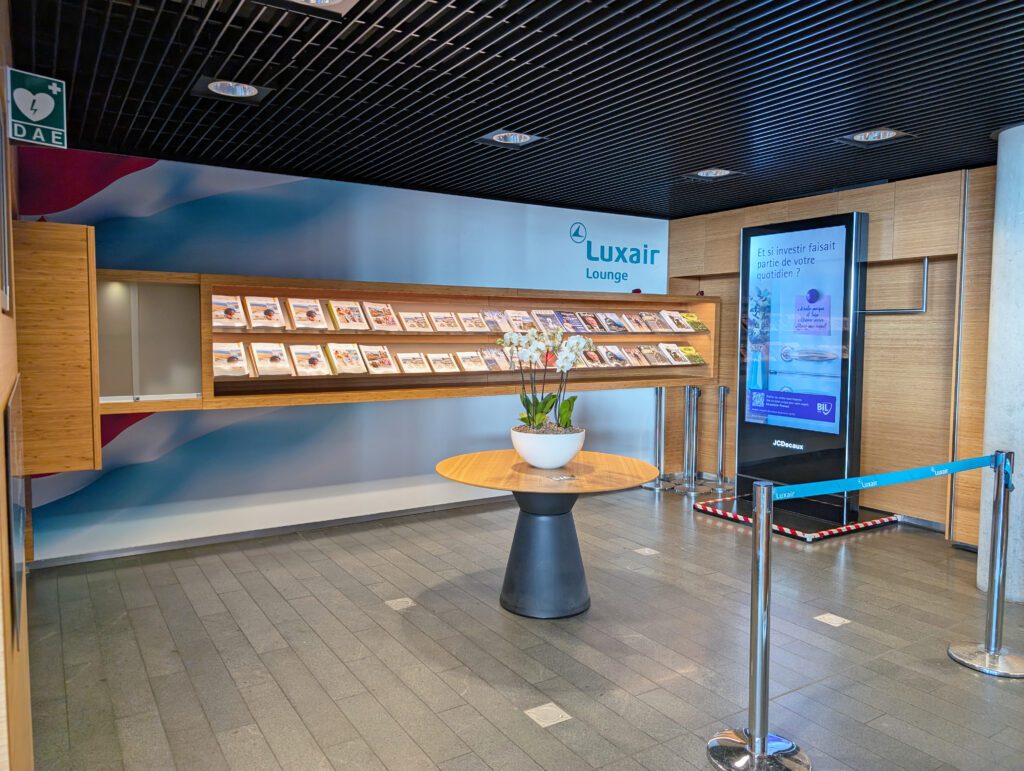 Eingangsbereich der Luxair Lounge mit einem runden Tisch mit einem weißen Blumentopf, einer digitalen Anzeige, einem Zeitschriftenständer und gefliestem Boden.