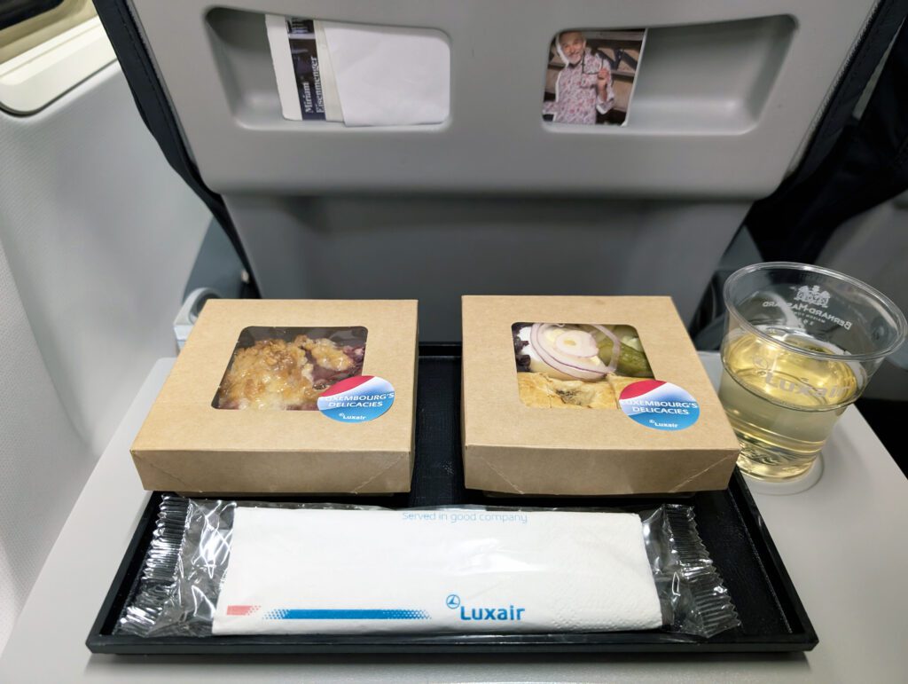 Ein Essenstablett in einem Flugzeug mit zwei abgepackten Mahlzeiten, einem Glas Weißwein, einer verpackten Serviette mit der Aufschrift „Luxair“ und einer Zeitschriftentasche im Hintergrund.