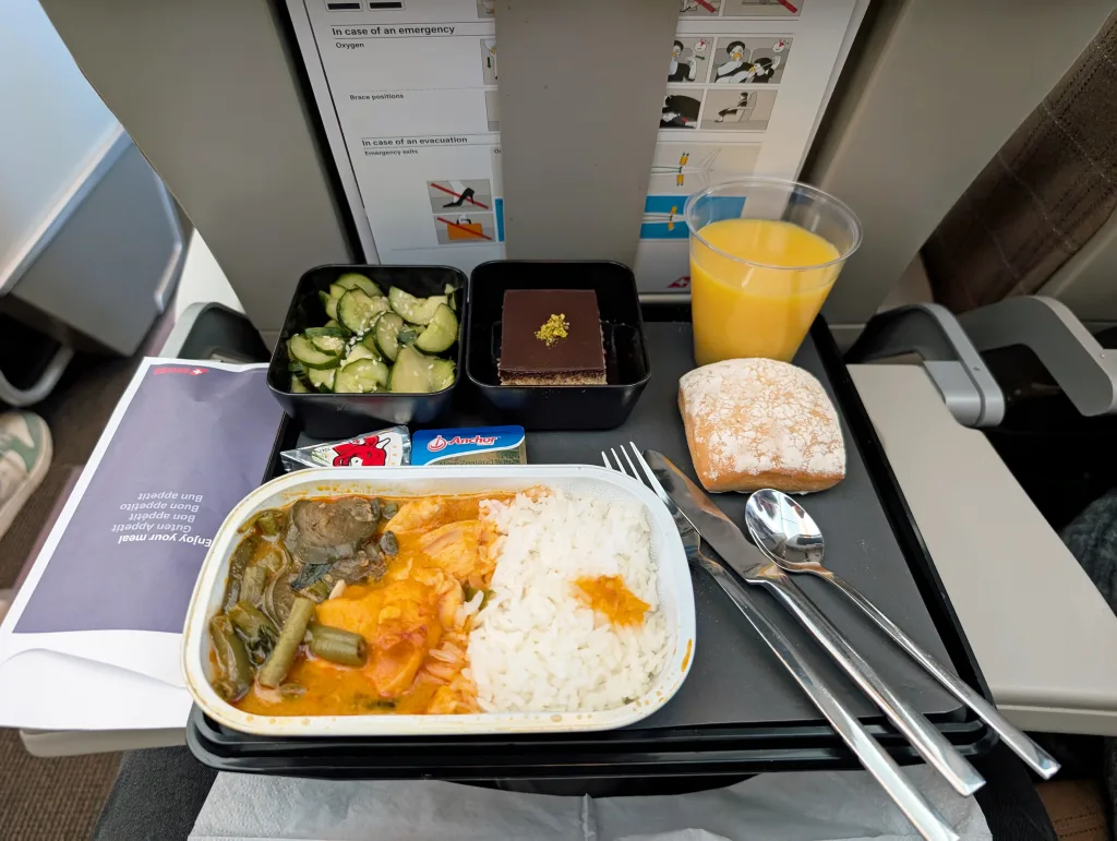 Airplane tray meal with rice, curry, vegetables, dessert, bread roll, butter, cucumber salad, and a cup of orange juice on a foldable tray table.