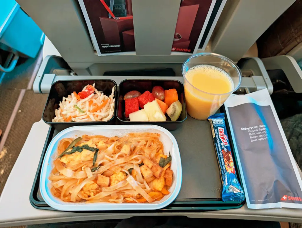 An airline meal on a tray with noodles, vegetable salad, fruit, a granola bar, a cup of orange juice, and a packaged napkin on an airplane tray table.