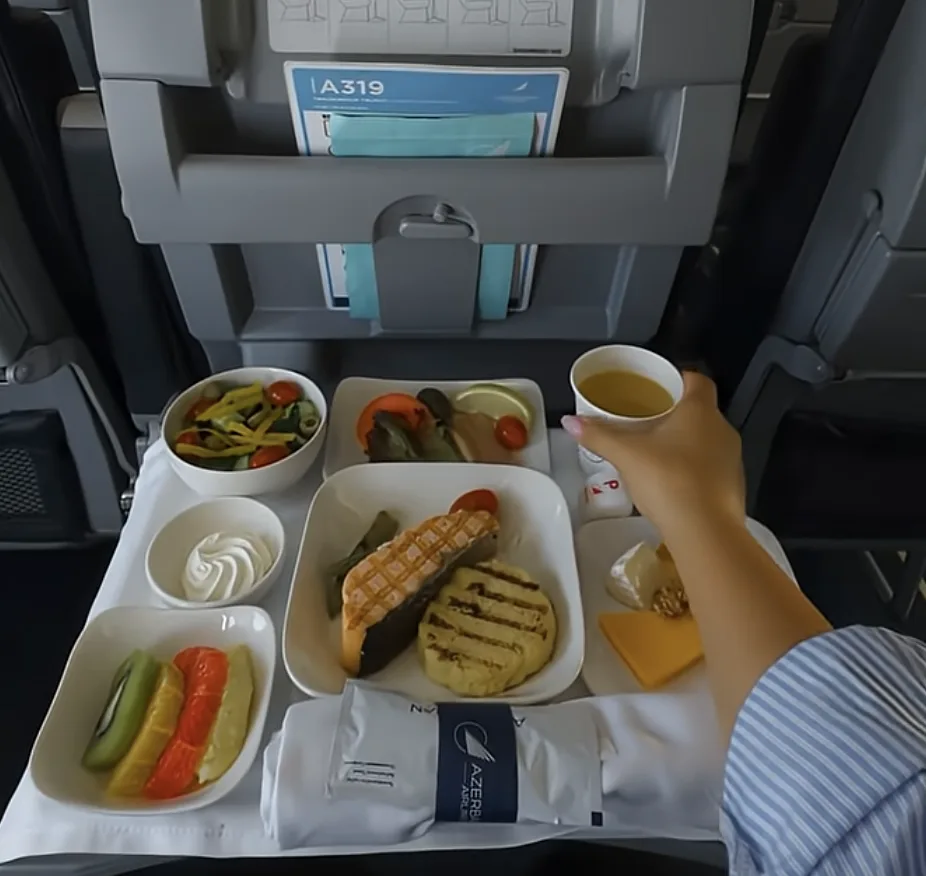 A person holds a cup of Southwest coffee above a tray of airline food, including bread, grilled chicken, vegetables, salad, cheese, dessert, and water, on an airplane seat tray table.