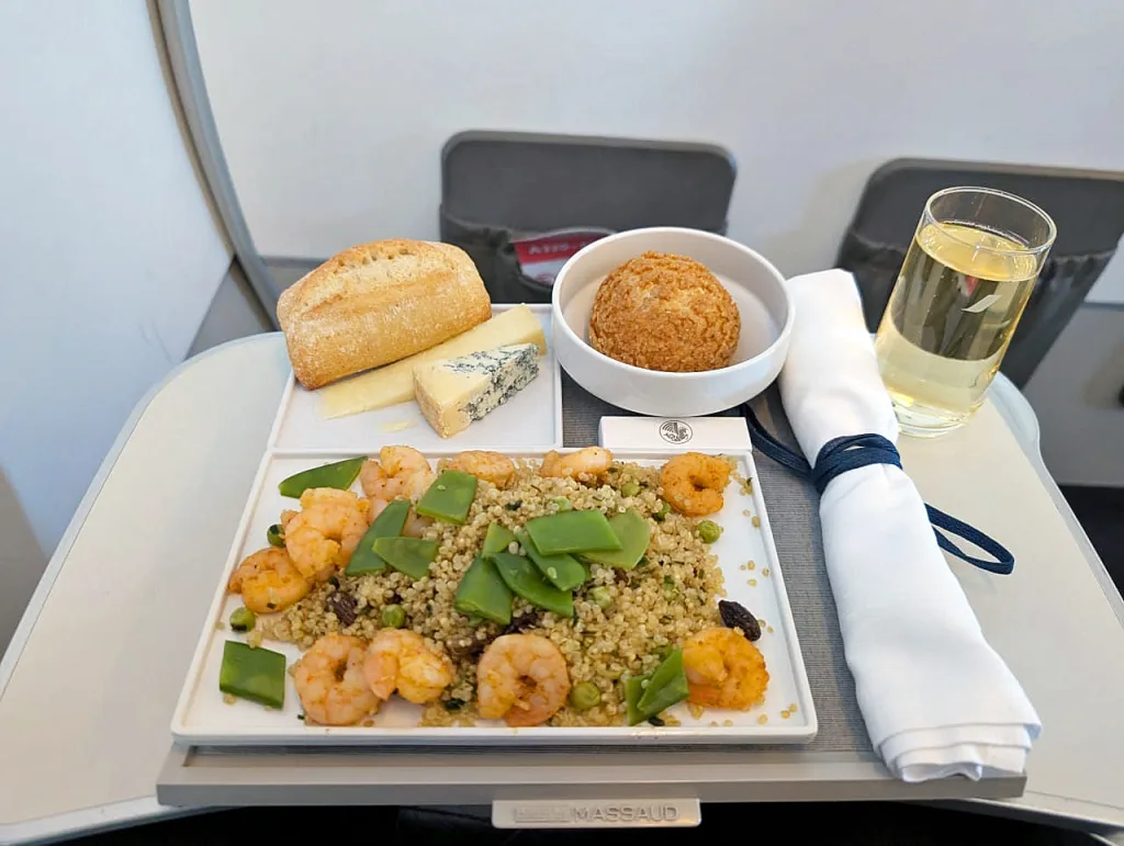 Air France: When Michelin Stars Meet High Altitude 7 A tray of Luxair food featuring shrimp and quinoa salad, a bread roll, cheese, dessert, and a glass of white wine on an airplane tray table invites a delightful Luxair review.