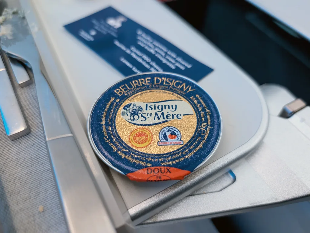 A round container of Isigny Sainte-Mère Beurre D’Isigny butter sits on a white tray next to a knife and a blue card, showcasing the premium quality often highlighted in Luxair food experiences.