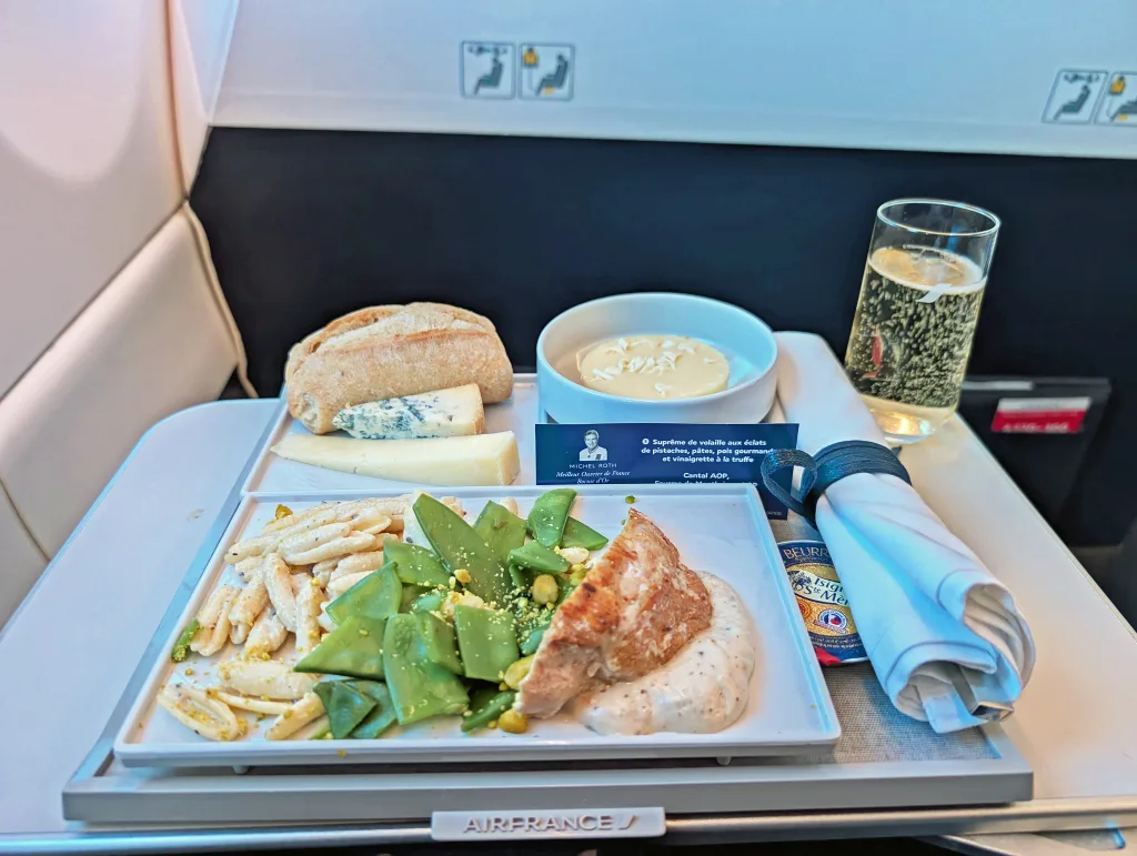 A tray of Luxair food includes pasta, green beans, chicken with sauce, bread, cheese, dessert, a glass of sparkling wine, and a rolled napkin—perfect for anyone seeking an authentic Luxair review of their in-flight dining experience.