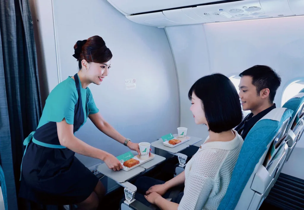 A flight attendant serves meals to two seated passengers on an airplane. Each tray has a cup of Southwest coffee and a pastry. The passengers appear attentive and engaged with the attendant.