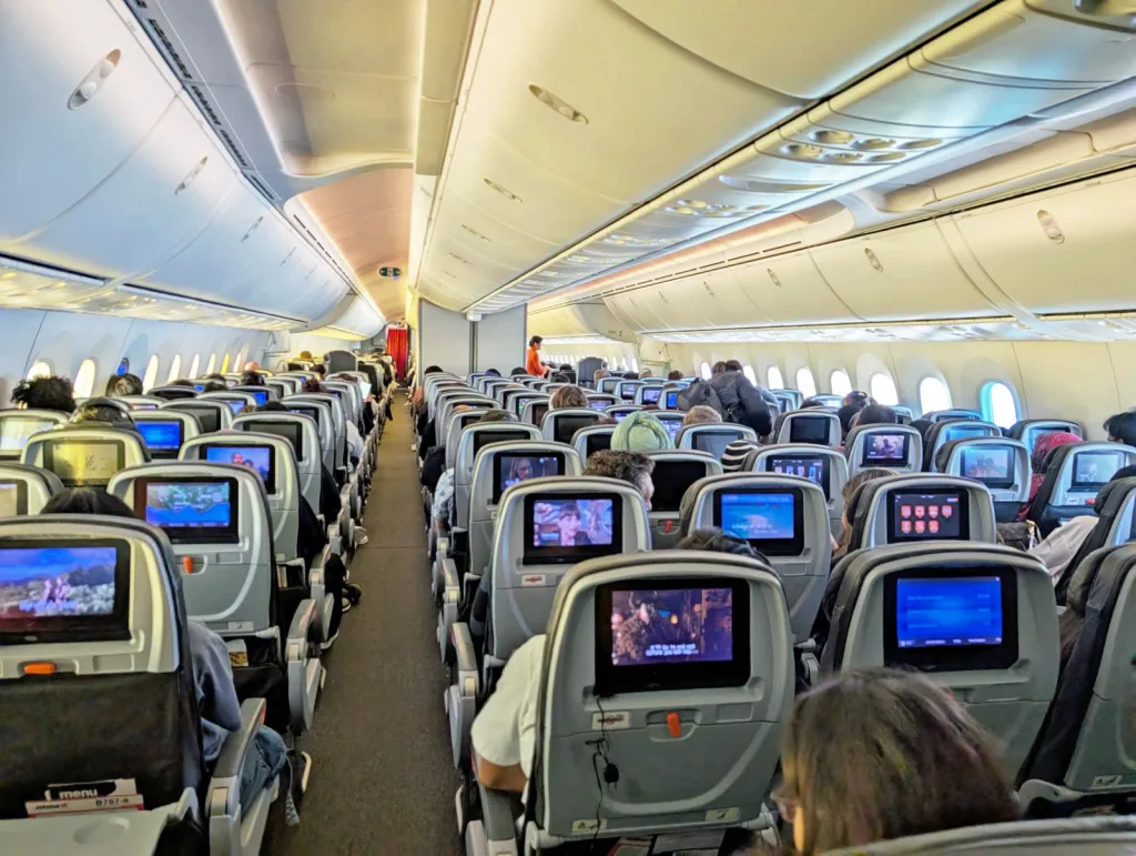Airplane economy cabin with rows of passengers seated, seatback entertainment screens on, and overhead bins closed.