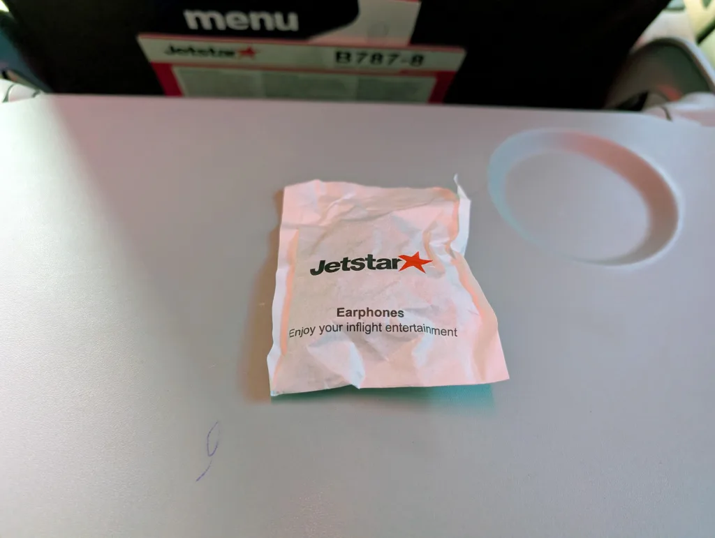 A crumpled Jetstar earphones packet sits on an airplane tray table, with a seat back and menu visible in the background.