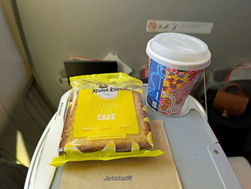 A packet of lemon tea cake and a colorful paper cup with a lid are on an airplane tray table with the Jetstar logo visible.