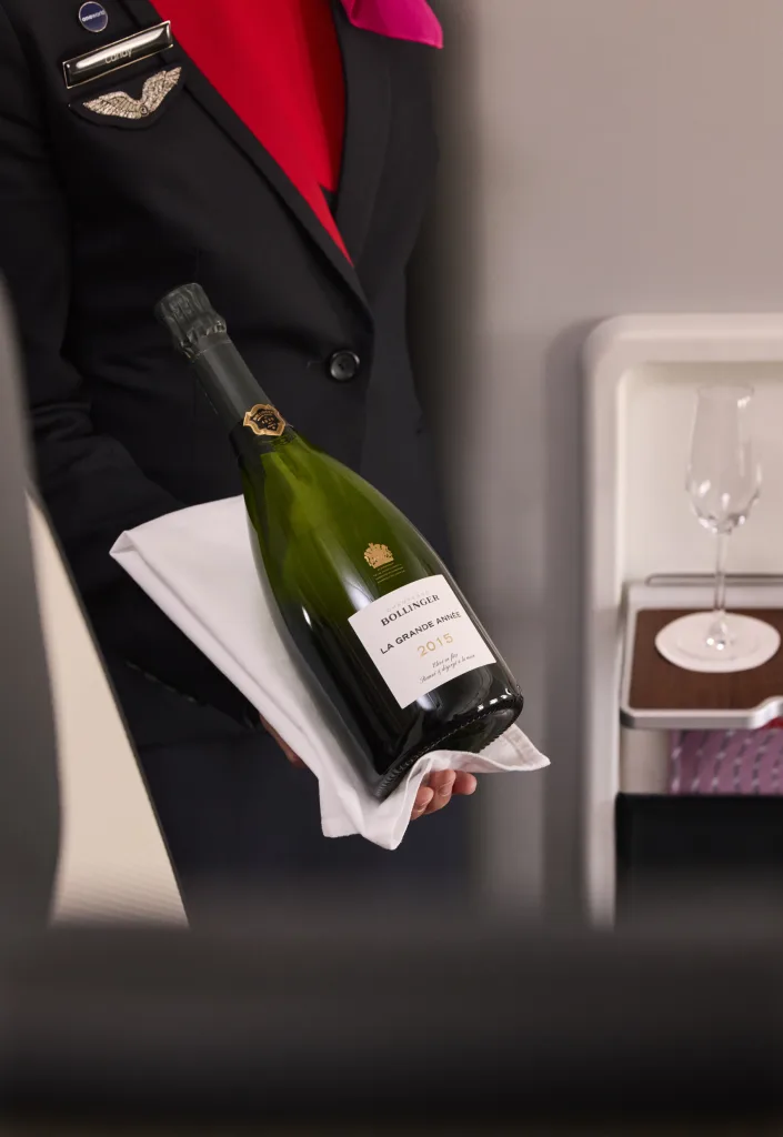 A Korean Air flight attendant in uniform presents a bottle of Bollinger La Grande Année 2015 champagne on a white cloth beside an empty glass, highlighting the airline’s commitment to sustainability with plant-based containers.