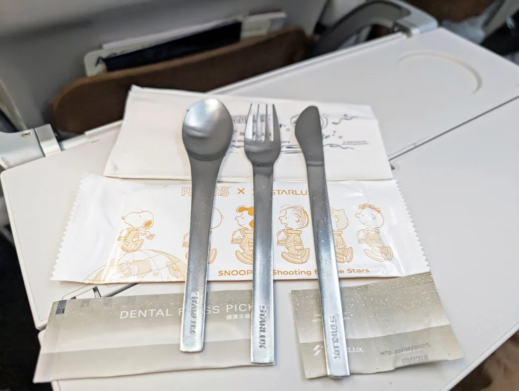 A set of metal spoon, fork, and knife is arranged on an airplane tray table, along with napkins, a dental floss pick, and a Snoopy-themed packaged item.