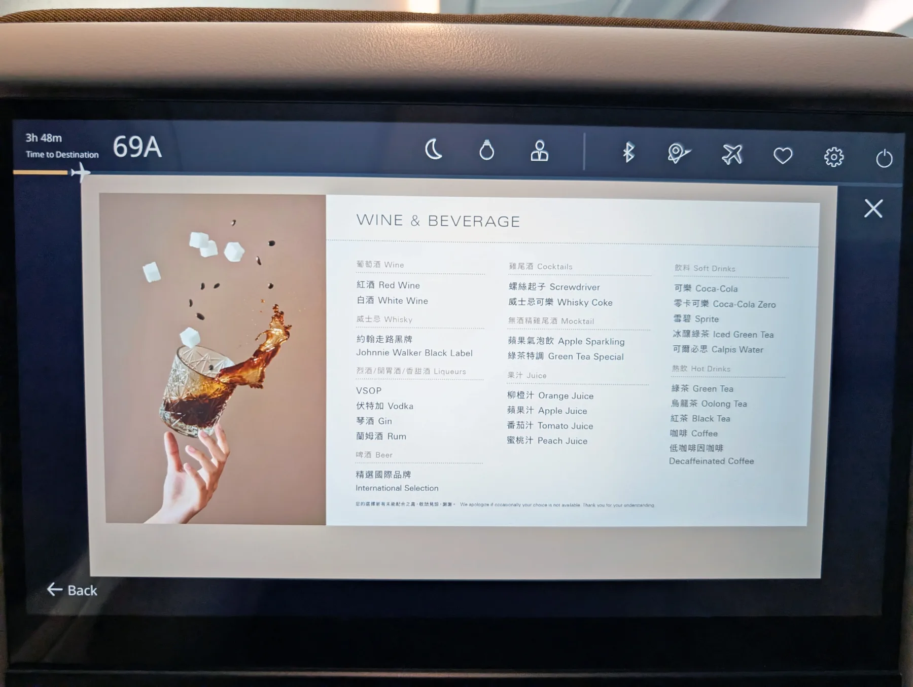 An in-flight entertainment screen displays a menu of wine, spirits, cocktails, soft drinks, juices, and hot beverages next to an image of a hand holding a glass with a splash and sugar cubes.