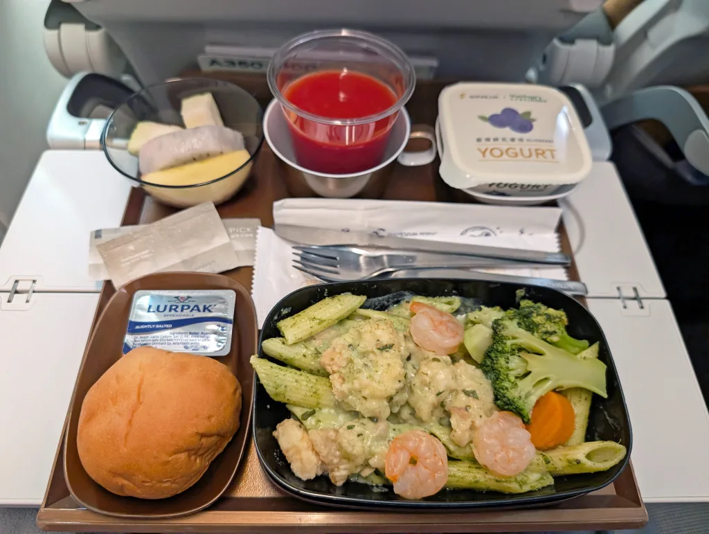 Airline meal tray with pasta, shrimp, broccoli, carrot, bread roll with butter, fruit slices, yogurt, red drink, and utensils placed on an airplane seat tray table.