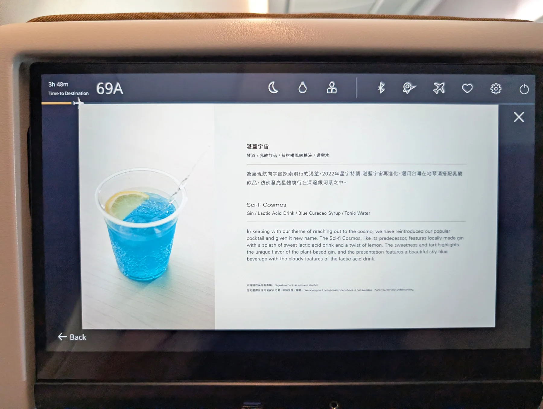 A plastic cup with a blue drink and a lemon slice sits on a tray table, shown on an airplane seatback screen displaying a description of the beverage.
