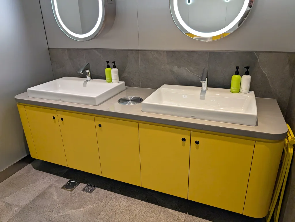 Two rectangular sinks with faucets are set in a yellow vanity with gray countertop, under two round mirrors in a modern bathroom. Soap dispensers are placed beside each sink.