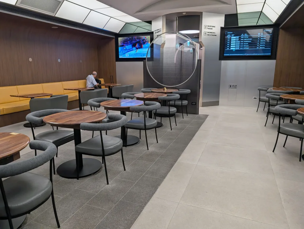 Modern airport lounge with empty tables and chairs, one person seated at the back, wall-mounted screens displaying sports and flight information.