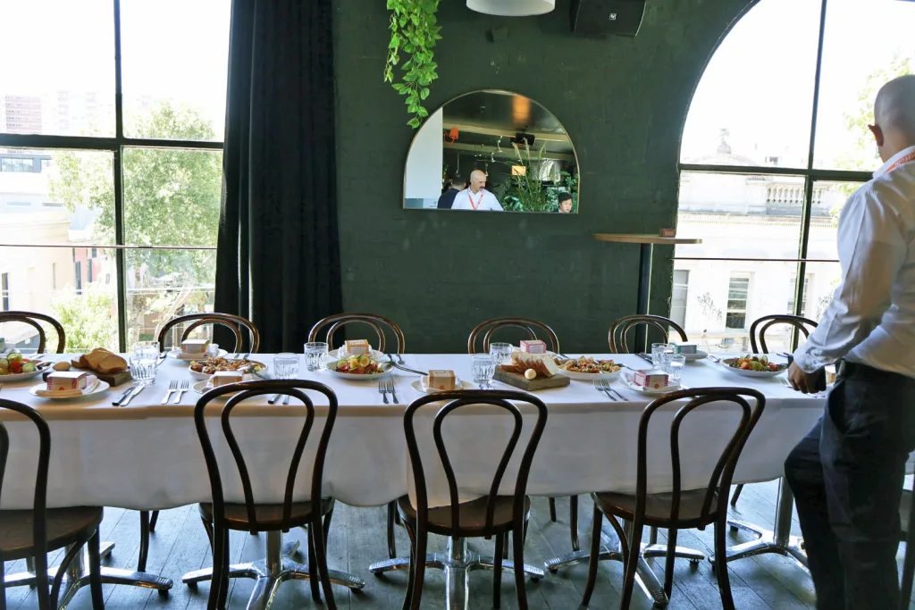 A long dining table set with plates, cutlery, and food in a restaurant; a person arranges items at one end, while another—dressed in huntsman tailoring—is visible in a mirror on the wall.