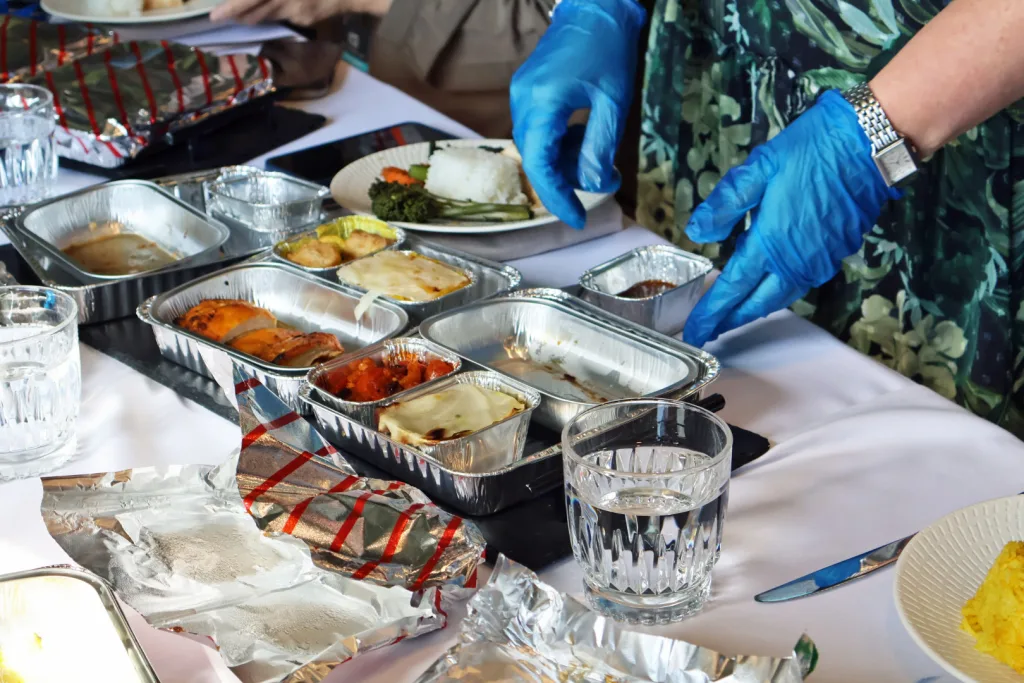 Wearing blue gloves, a person serves meals from foil trays onto plates on a table set with Starlux elegance—glasses of water and other dishes arranged with huntsman tailoring precision.