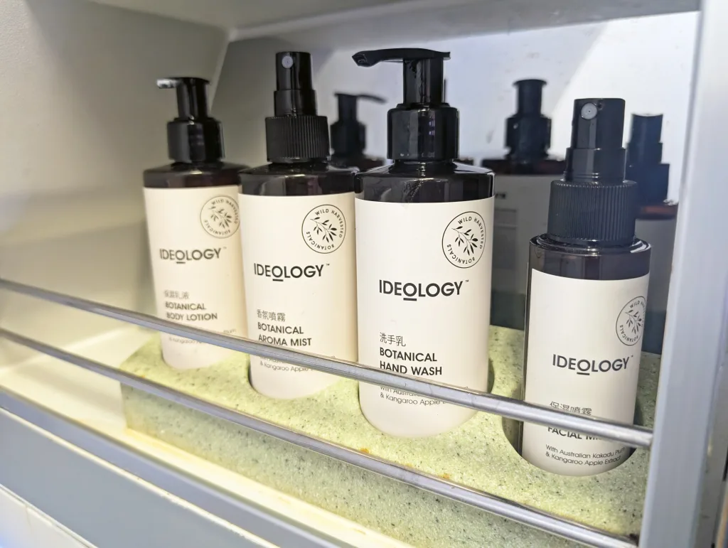 Four IDEOLOGY brand bottles labelled as botanical body lotion, aroma mist, hand wash, and facial wash are displayed on a shelf with a metal rail in front.
