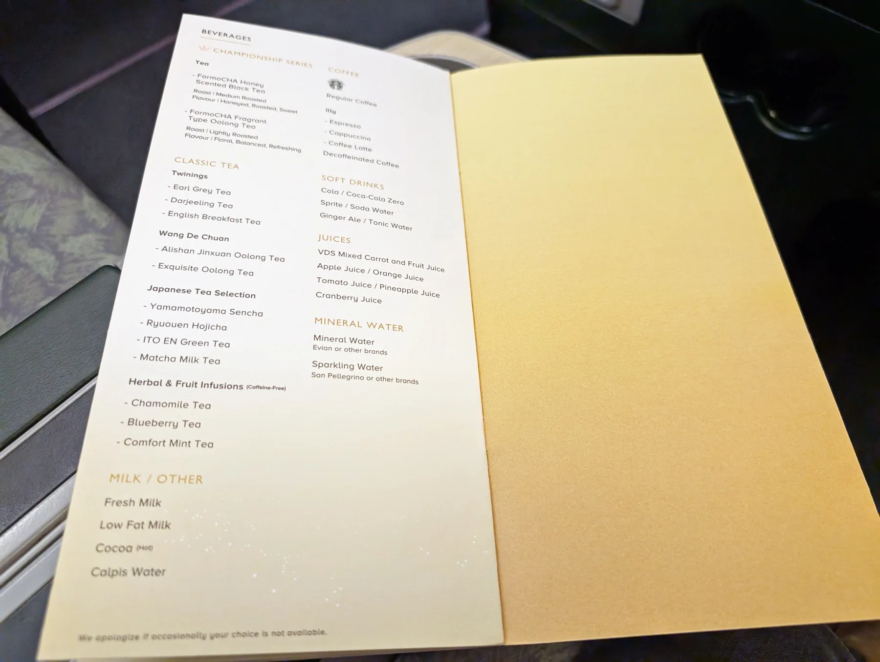 A close-up of an airline drinks menu on a tray table, listing tea, coffee, soft drinks, juices, milk, and water options. The right page is blank.