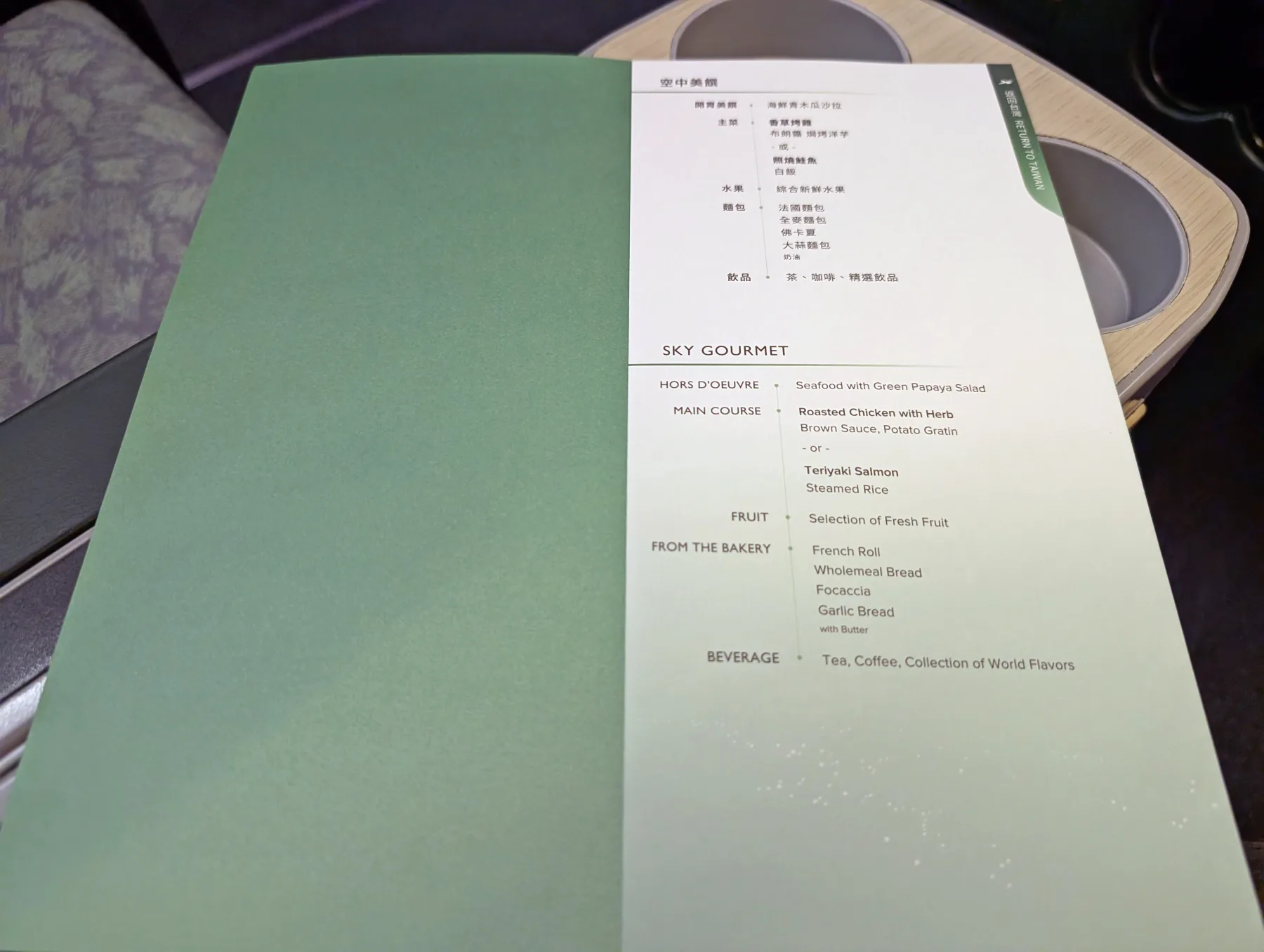 An airline menu on a tray table lists hors d’oeuvre, main course options, fruit, bakery items, and beverages in English and Chinese.