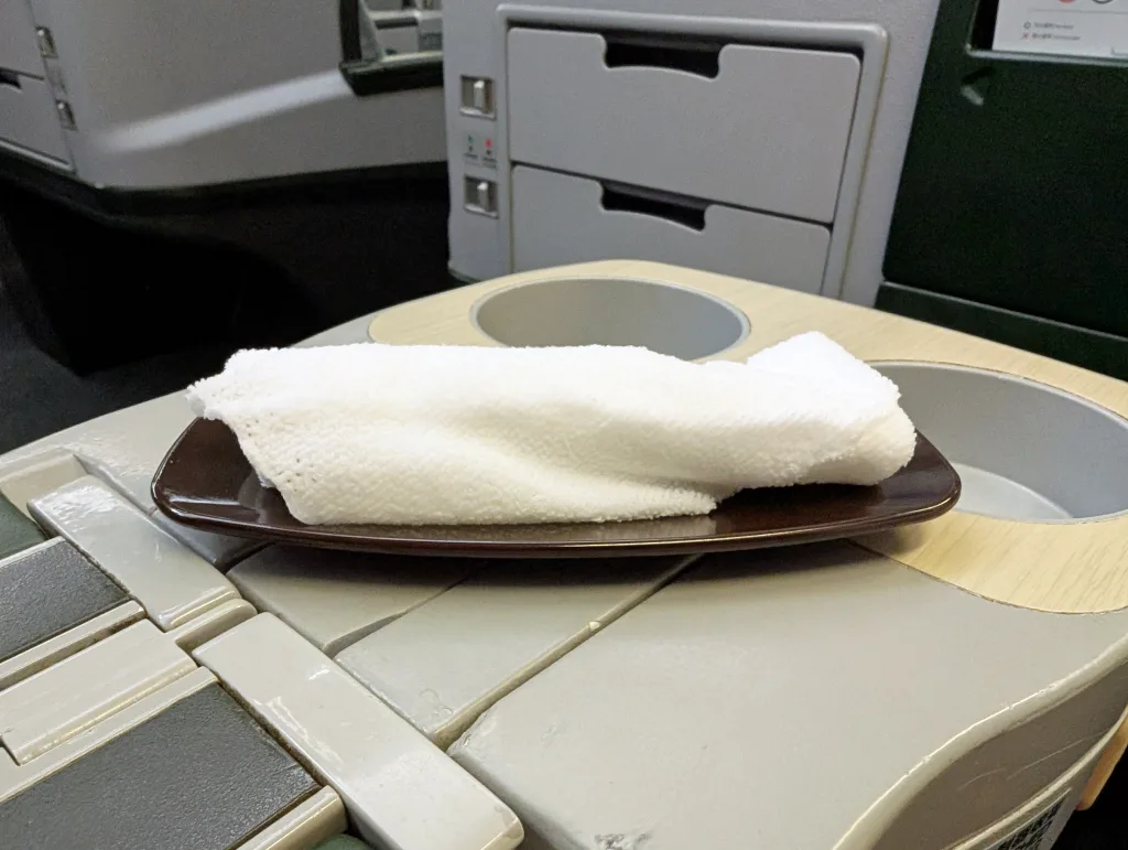 A rolled white towel on a dark tray is placed on an aeroplane seat's armrest with cup holders and a storage compartment visible.