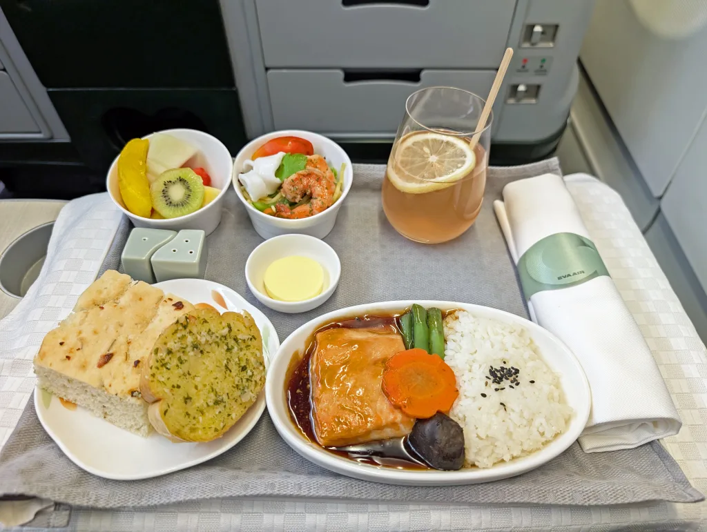 Airline meal tray with rice and fish main course, vegetables, bread, butter, salad, fruit, and a glass of iced tea with a lemon slice on a serviette-covered tray table.