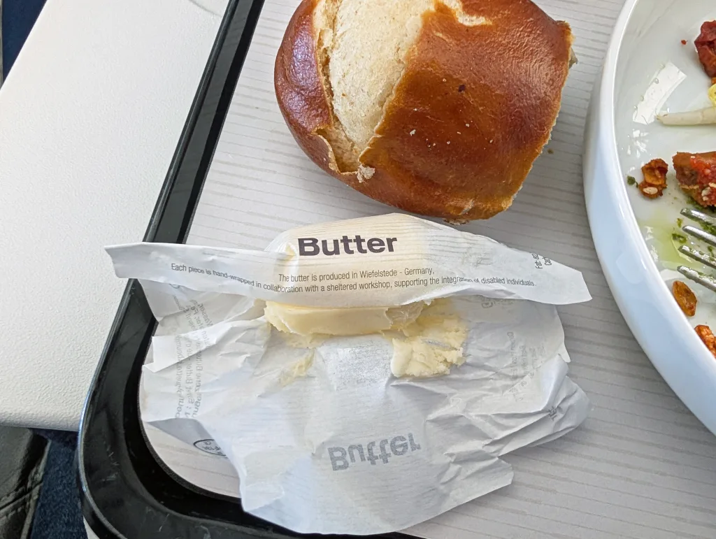 An opened butter packet with some butter remaining sits next to a bread roll on a tray, with part of a plate of food and cutlery visible.