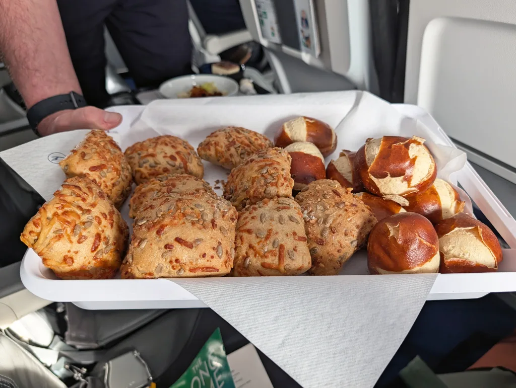 A tray with cheese-topped bread rolls and small pretzel rolls is being served on an aeroplane, with serviettes under the bread.