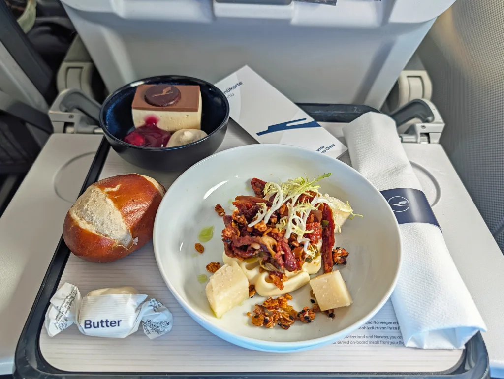 A tray of airline food with a bread roll, butter, dessert, serviette, and a main dish of sliced meat, cheese cubes, and garnish, set on an aeroplane tray table.