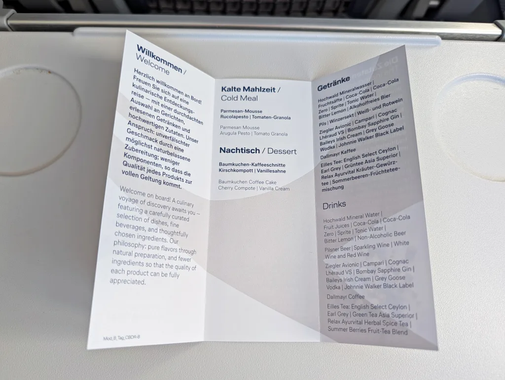An airline meal menu displayed on a tray table, printed in German and English, listing options for cold meal, dessert, and drinks.