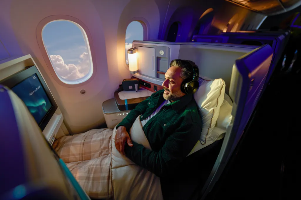 A man wearing headphones relaxes in a business class aeroplane seat, enjoying his Korean Air awards experience as he watches an in-flight screen with a blanket over his lap and daylight visible through the window.
