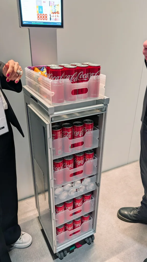 A drinks trolley stocked with Coca-Cola cans and snacks, reminiscent of Korean Air awards service, stands between two people—one in white trainers and the other in black.