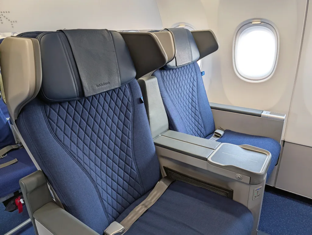 Two blue upholstered aeroplane seats with quilted patterns in a premium cabin, featuring wide armrests and seatback trays, next to windows.