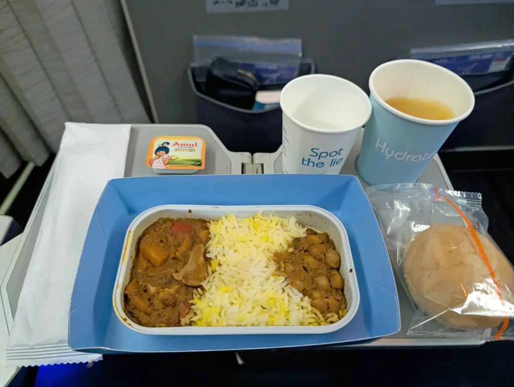 A tray with a meal of rice, curry, bread roll, butter, a cup of juice or tea, and a cup of water served on an aeroplane tray table.